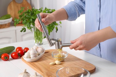 Photo of Woman squeezing garlic with press at white wooden table in kitchen, closeup