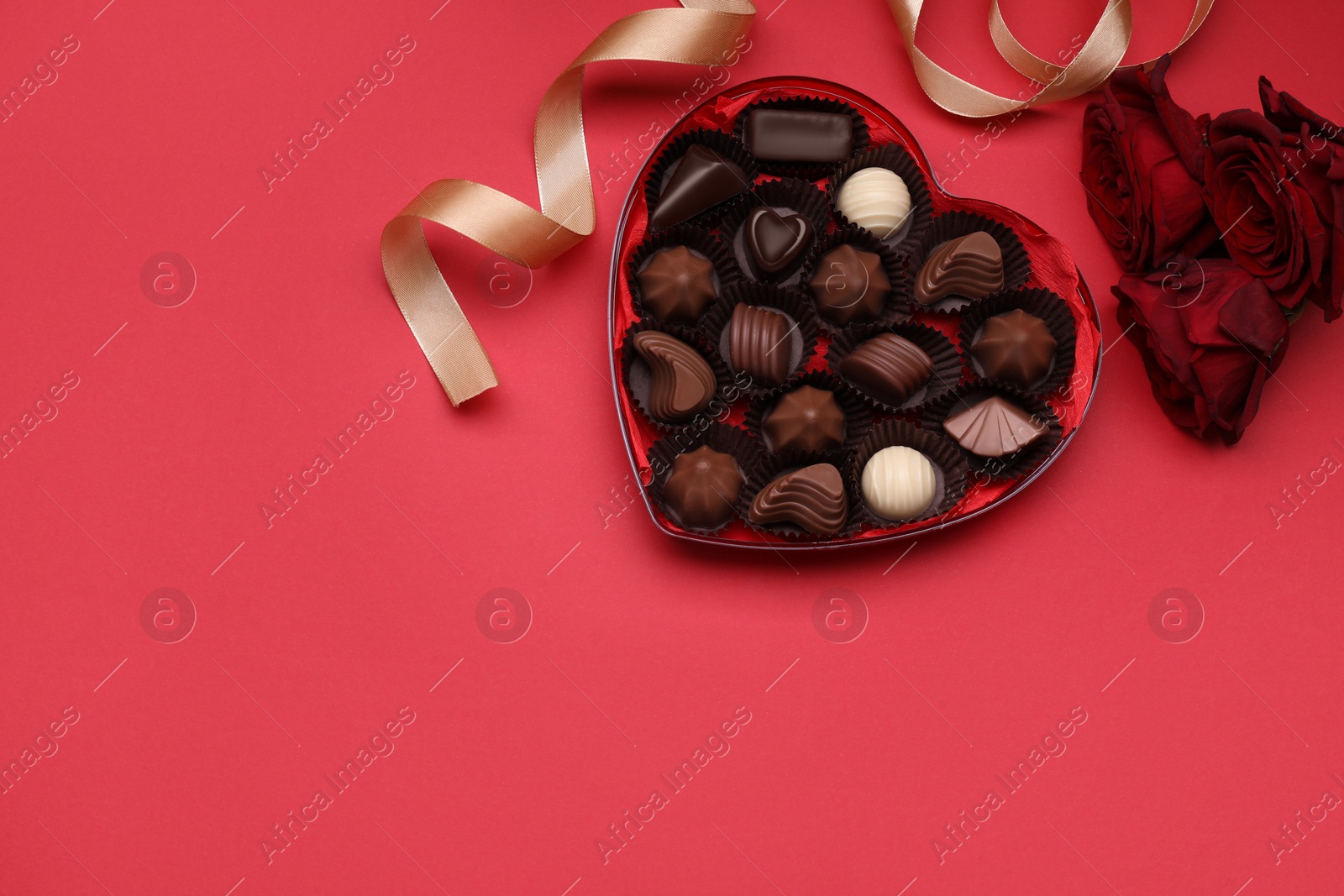 Photo of Heart shaped box with delicious chocolate candies, roses and ribbon on red background, flat lay. Space for text