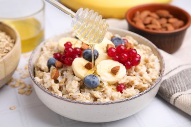 Honey pouring into bowl of oatmeal with berries, almonds and banana slices on white wooden table, closeup