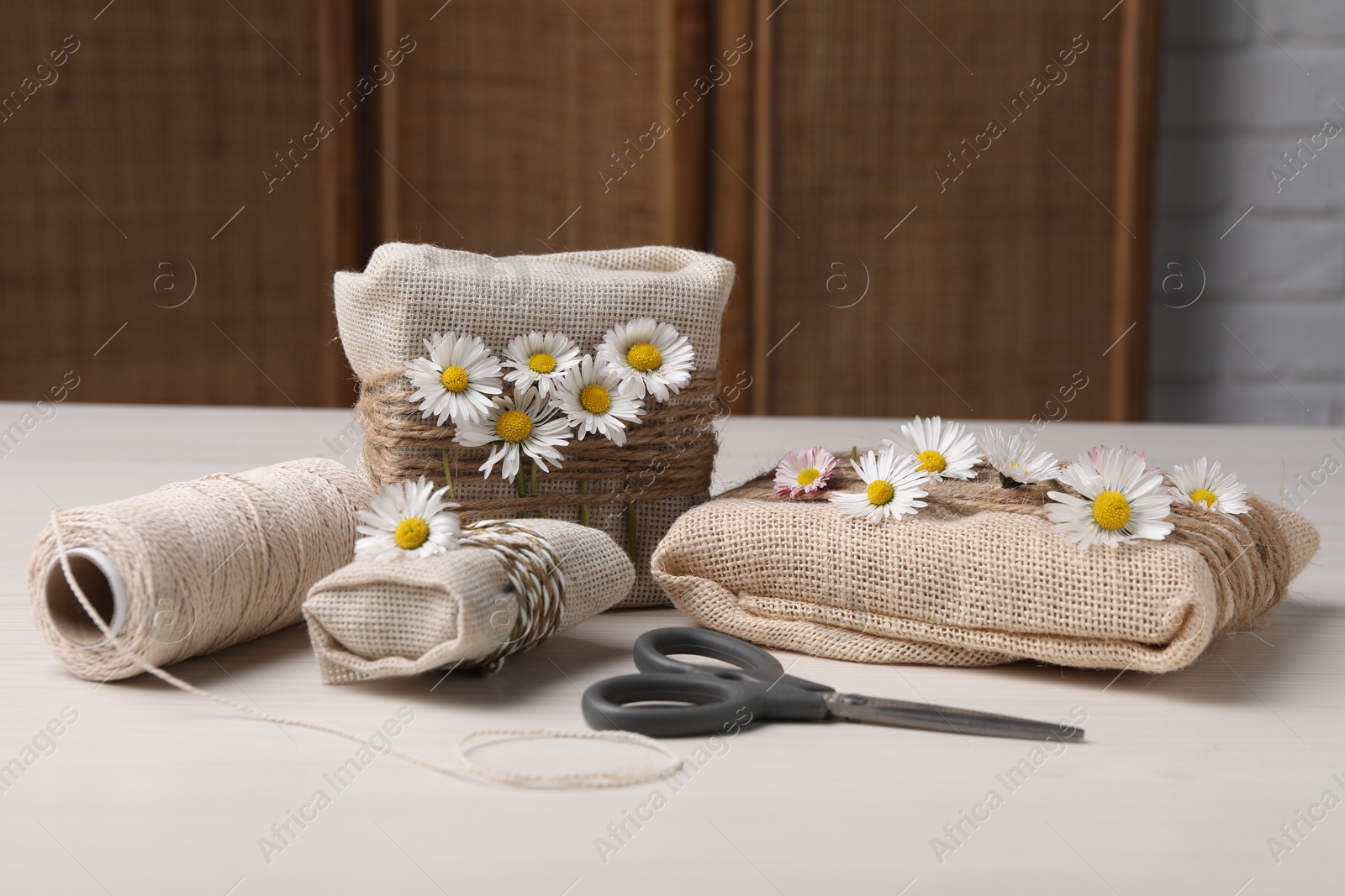 Photo of Gifts packed in burlap fabric, beautiful chamomiles, ropes, spool of threads and scissors on white table