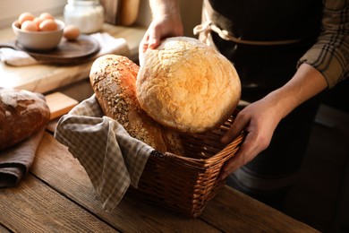 Photo of Man holding wicker basket with different types of bread at wooden table indoors, closeup