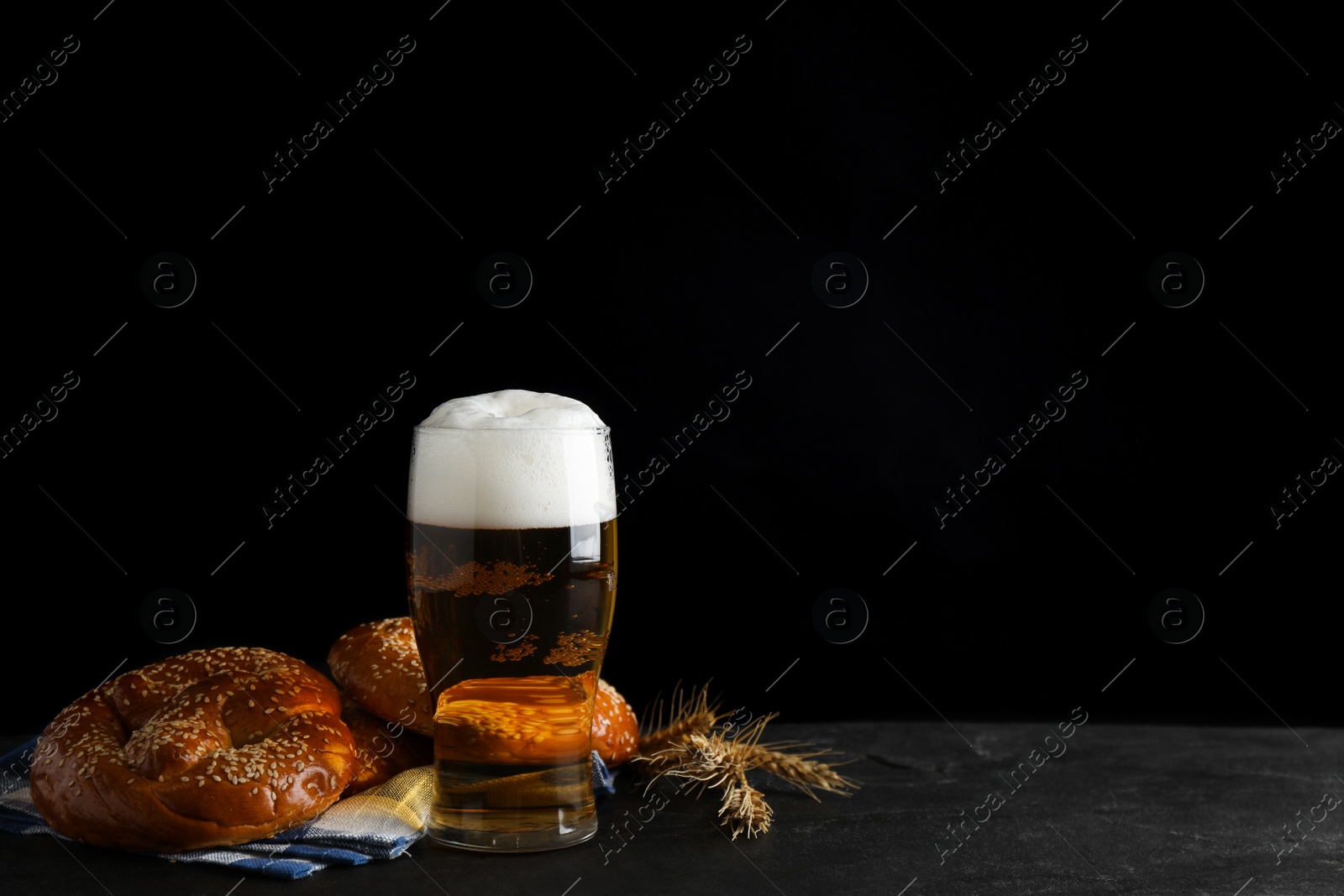 Photo of Tasty pretzels, glass of beer and wheat spikes on black table against dark background, space for text