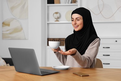 Photo of Muslim woman with cup of coffee using video chat on laptop at wooden table in room