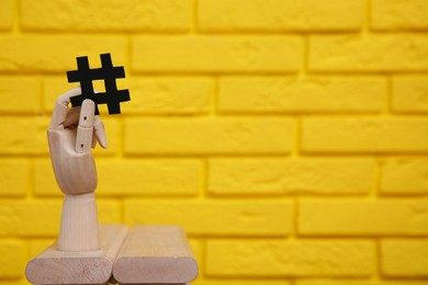 Wooden mannequin hand with paper hashtag symbol near yellow brick wall. Space for text
