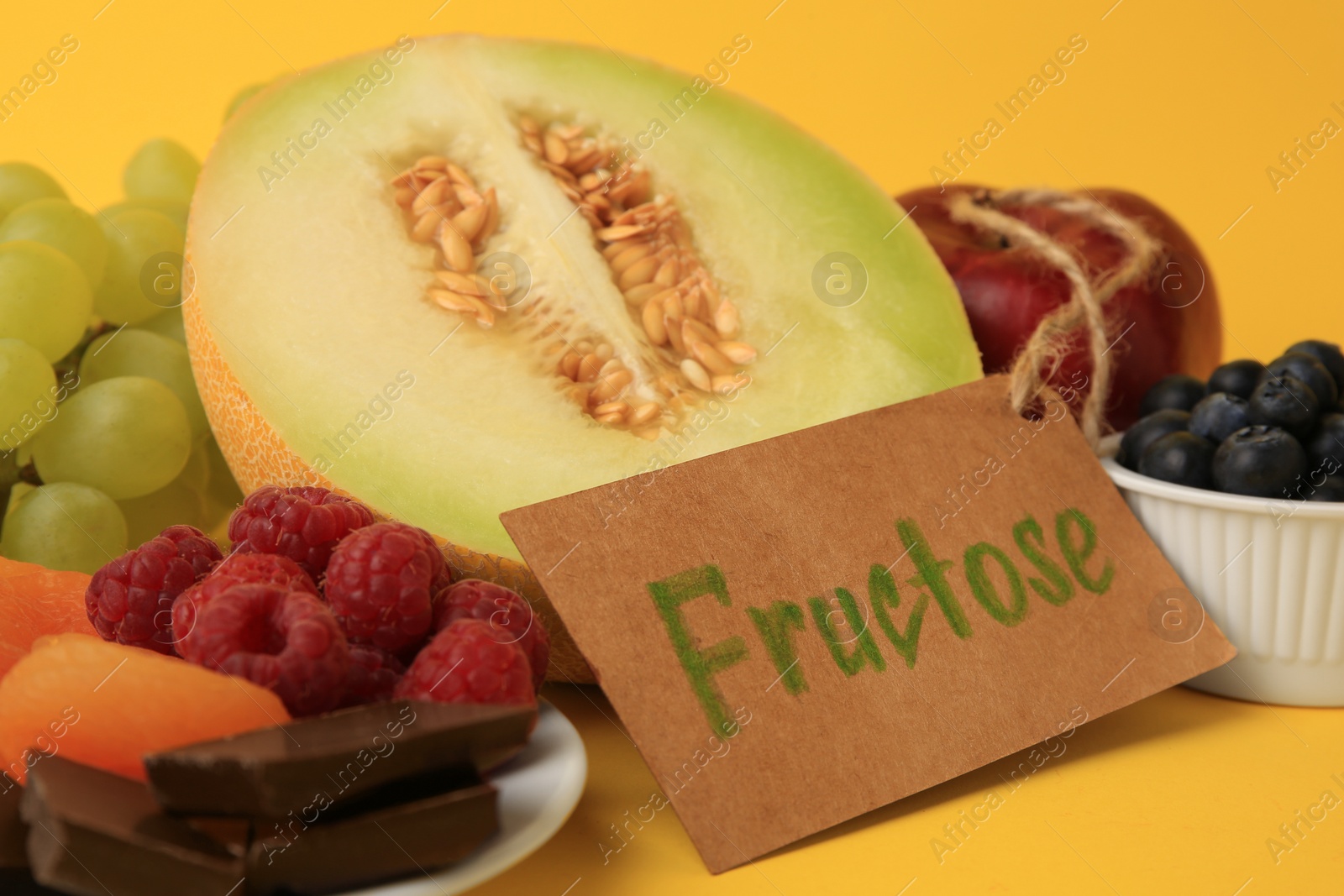 Photo of Label with word Fructose, delicious fruits, berries and pieces of chocolate on yellow background, closeup