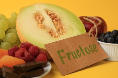 Photo of Label with word Fructose, delicious fruits, berries and pieces of chocolate on yellow background, closeup