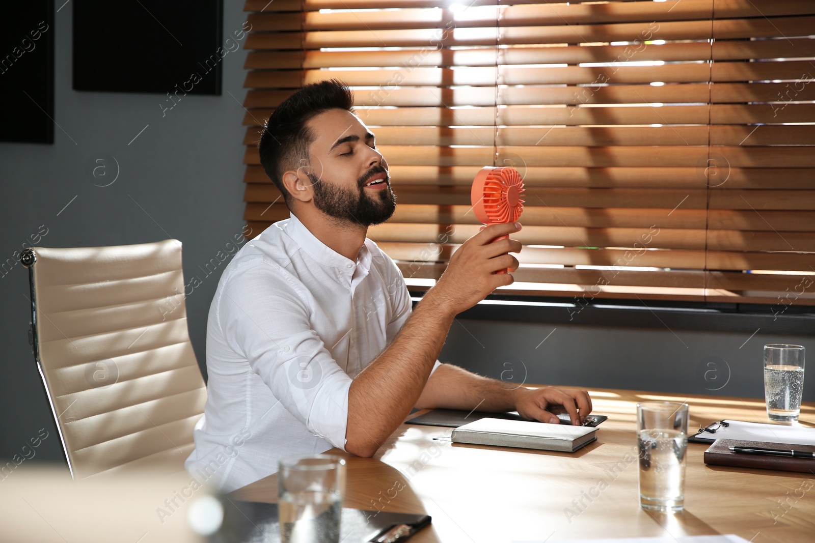 Photo of Man enjoying air flow from portable fan at workplace. Summer heat