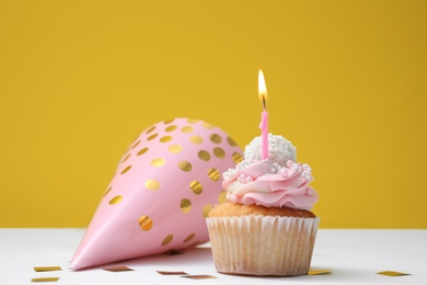 Delicious birthday cupcake with candle, party cap and confetti on white table against yellow background