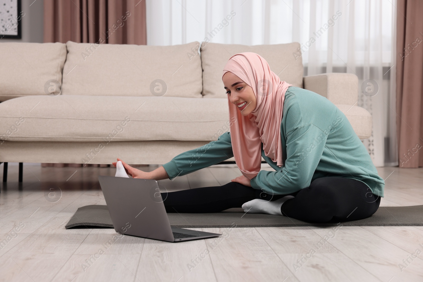 Photo of Muslim woman in hijab stretching near laptop on fitness mat at home