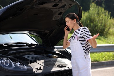 Stressed woman talking on smartphone while looking under hood of broken car outdoors