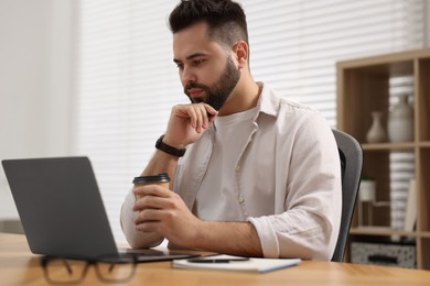 Young man with cup of coffee watching webinar at table in room