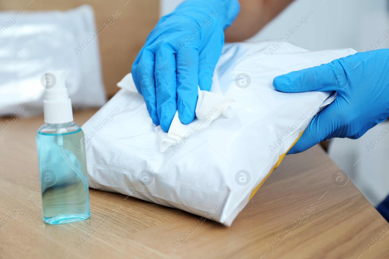 Photo of Woman cleaning parcel with wet wipe and antibacterial spray at wooden table, closeup. Preventive measure during COVID-19 pandemic