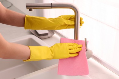 Photo of Woman in gloves cleaning faucet of kitchen sink with rag, closeup
