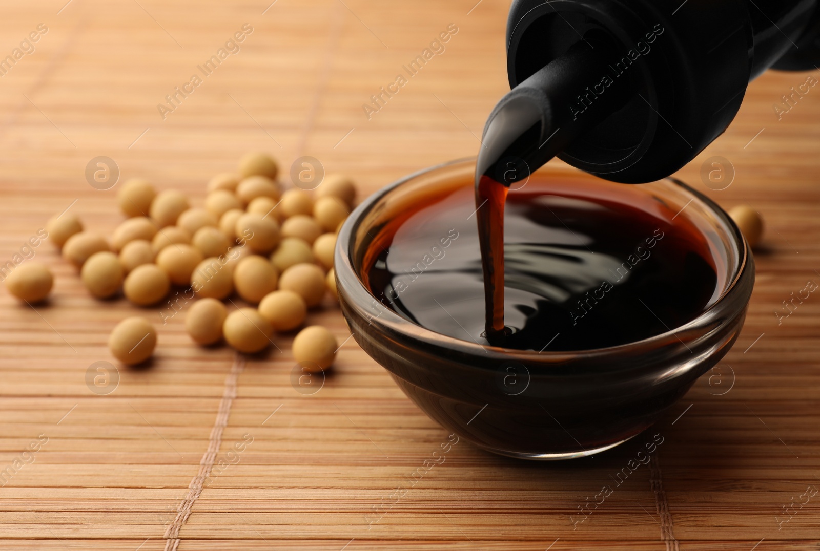 Photo of Pouring soy sauce from bottle into bowl and soybeans on bamboo mat, closeup