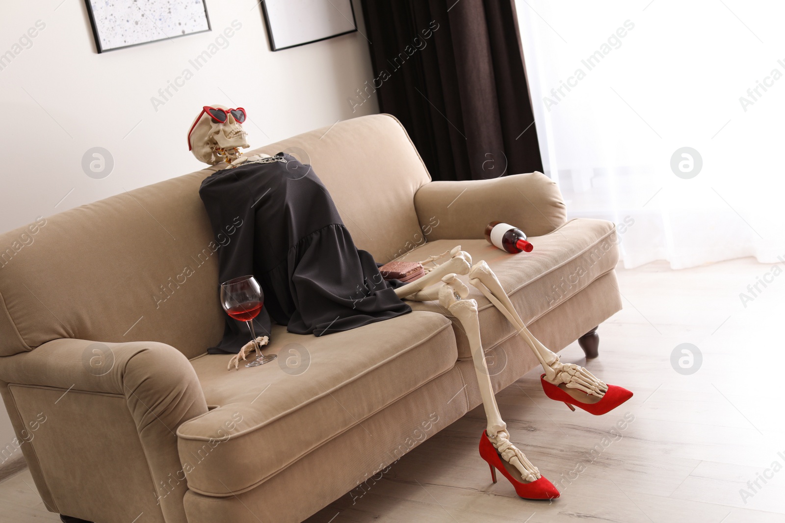 Photo of Skeleton in dress with wine sitting on sofa at home