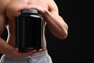 Young man with muscular body holding jar of protein powder on black background, closeup. Space for text