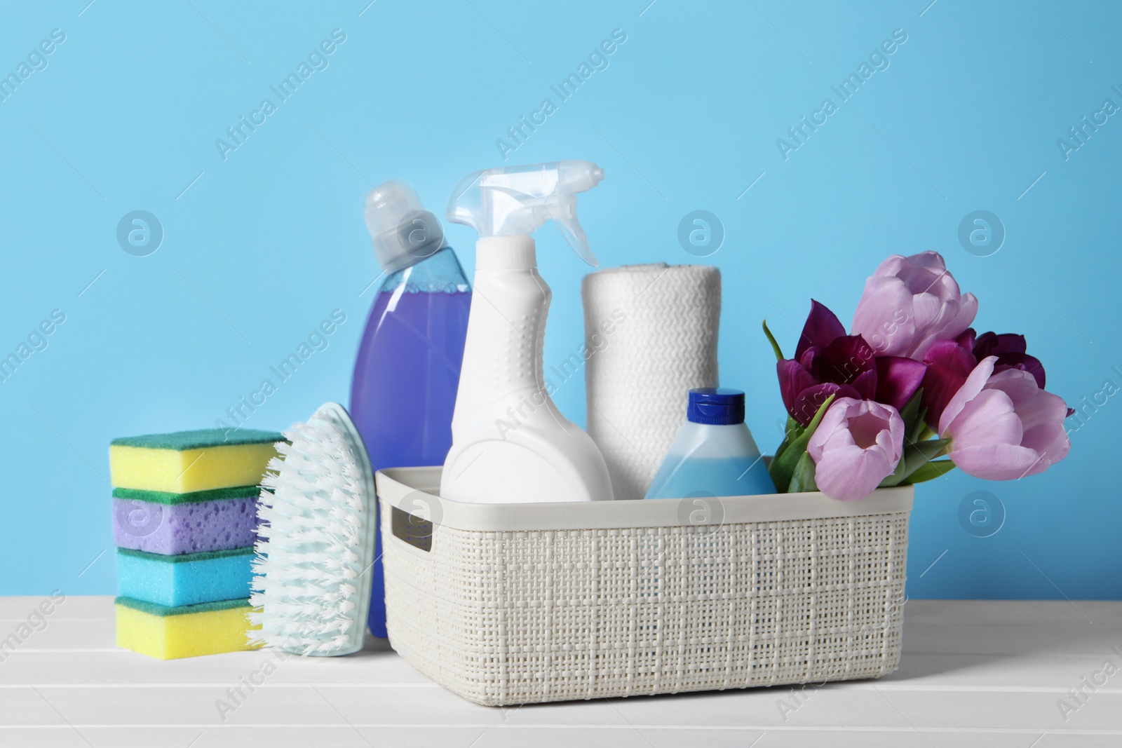 Photo of Spring cleaning. Basket with detergents, flowers and tools on white wooden table against light blue background