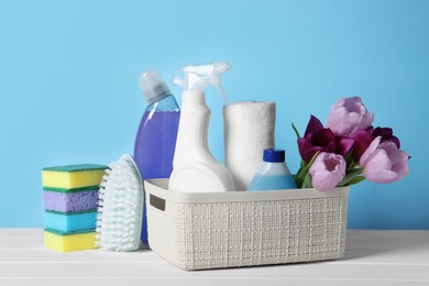 Photo of Spring cleaning. Basket with detergents, flowers and tools on white wooden table against light blue background