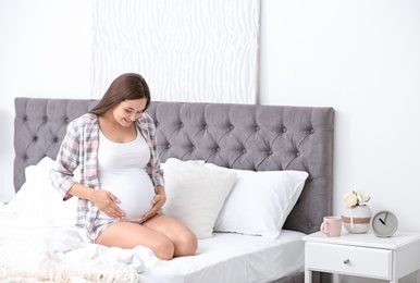 Photo of Young beautiful pregnant woman sitting on bed and touching her belly at home