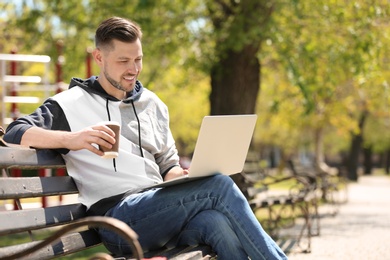 Portrait of young man with laptop and cup of coffee on bench in park