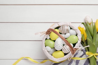 Photo of Easter eggs in basket and tulips on white wooden table, flat lay. Space for text