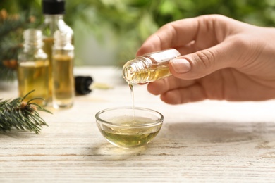 Photo of Woman pouring conifer essential oil into bowl on white wooden table, closeup