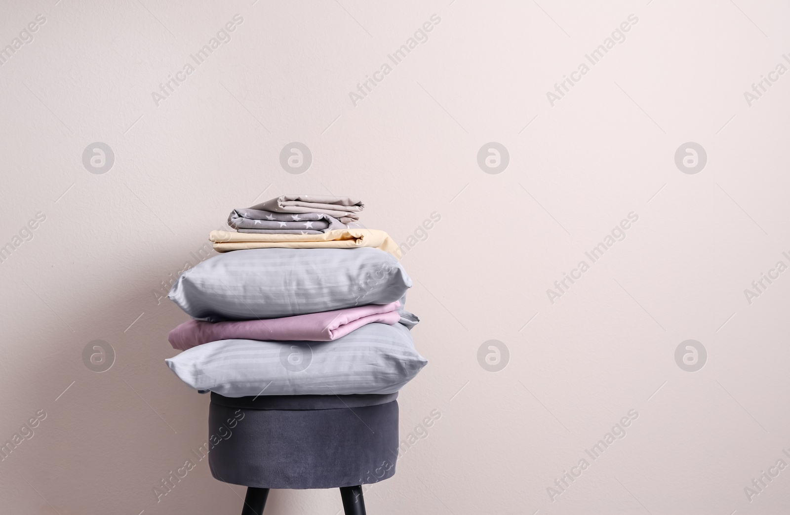 Photo of Stack of clean bed sheets and pillows on stool near beige wall. Space for text