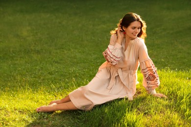 Photo of Beautiful woman in embroidered dress sitting on green grass outdoors, space for text. Ukrainian national clothes
