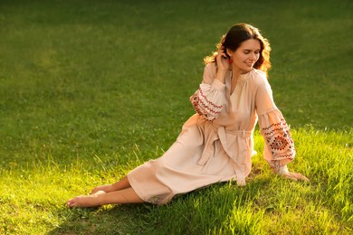 Beautiful woman in embroidered dress sitting on green grass outdoors, space for text. Ukrainian national clothes