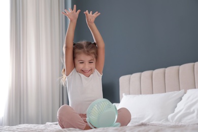 Photo of Little girl enjoying air flow from portable fan on bed in room. Summer heat