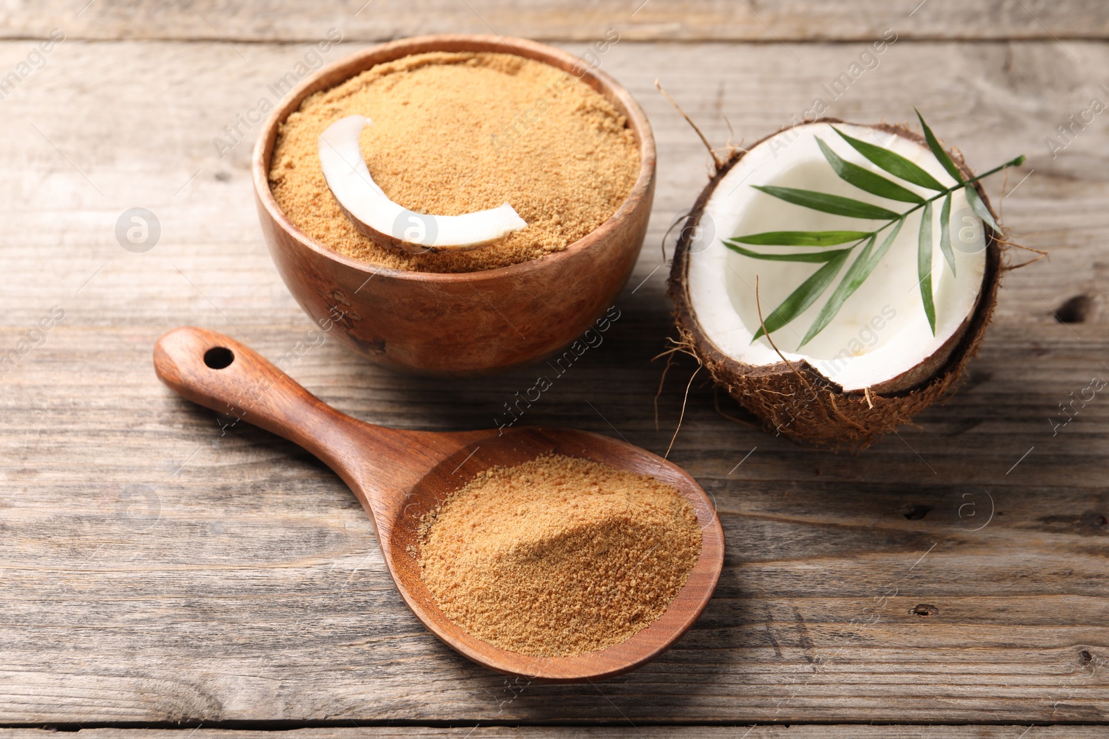 Photo of Coconut sugar in bowl, spoon, fruit and palm leaves on wooden table, closeup