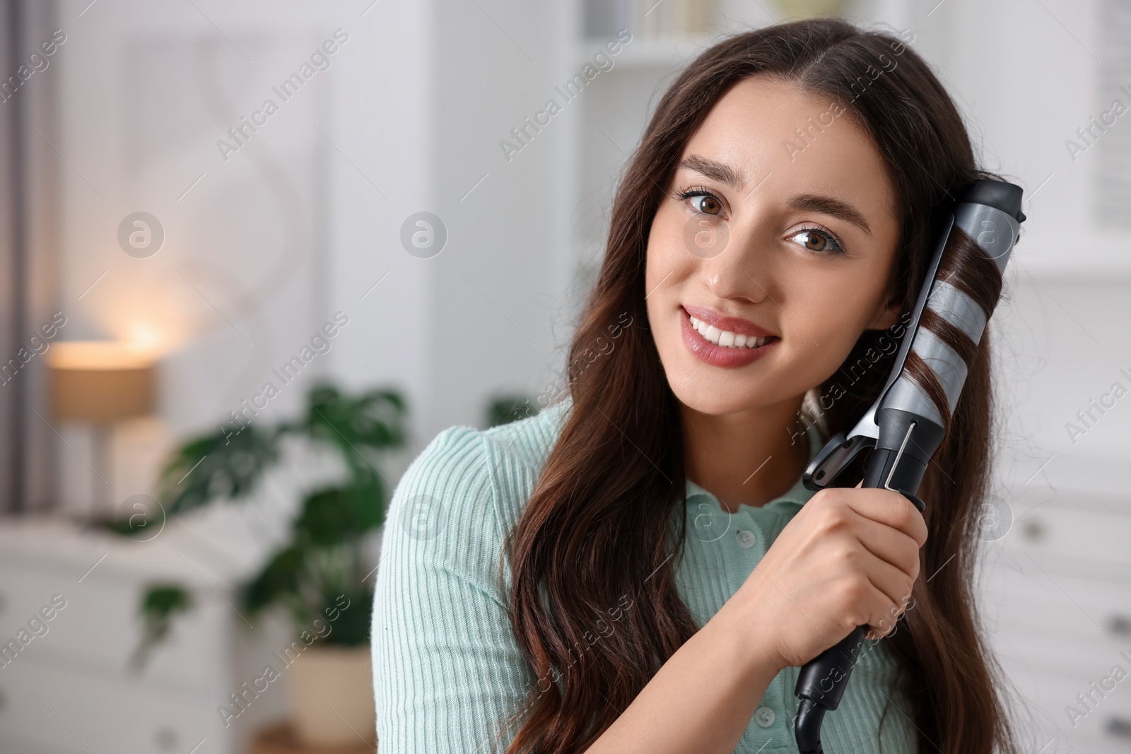 Photo of Smiling woman using curling hair iron at home. Space for text