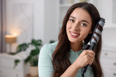 Photo of Smiling woman using curling hair iron at home. Space for text