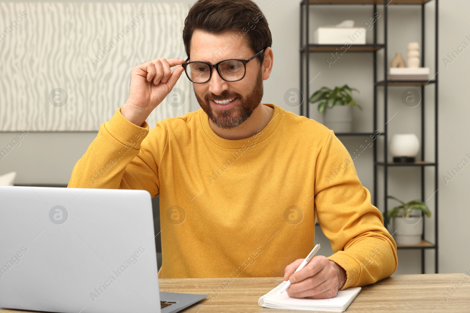 Photo of Man using laptop and writing something in notebook at wooden table indoors