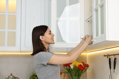 Photo of Beautiful young woman taking plate from kitchen cabinet at home