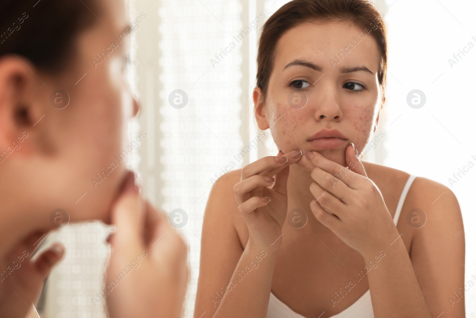 Photo of Teen girl with acne problem squeezing pimple near mirror in bathroom