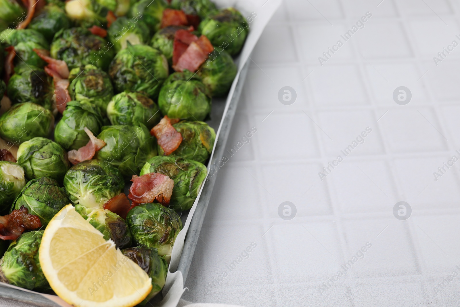 Photo of Delicious roasted Brussels sprouts, bacon and lemon in baking dish on white tiled table, closeup. Space for text