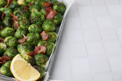 Photo of Delicious roasted Brussels sprouts, bacon and lemon in baking dish on white tiled table, closeup. Space for text