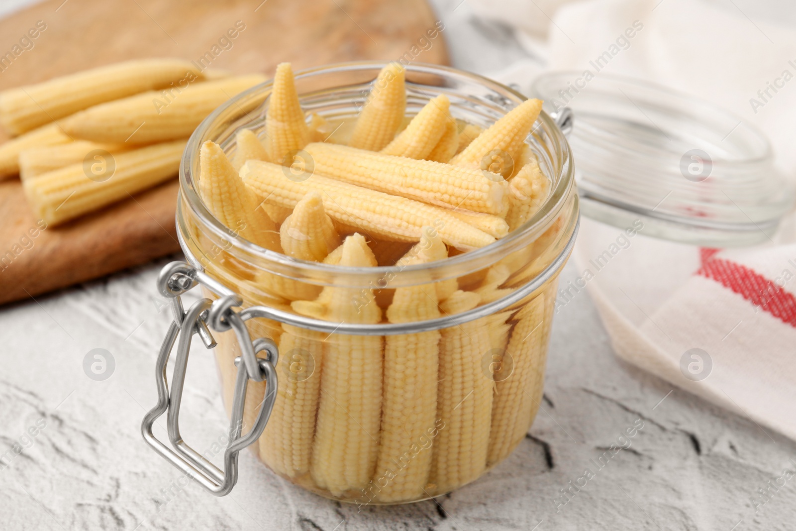 Photo of Jar of pickled baby corn on white textured table, closeup