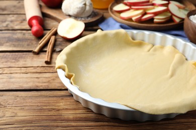 Photo of Baking dish with raw dough for apple pie and ingredients on wooden table