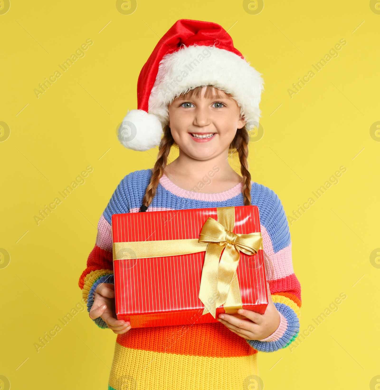 Photo of Cute little girl in Santa hat with Christmas gift on yellow background