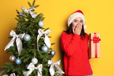 Beautiful young woman in Santa hat with gift box near Christmas tree on color background