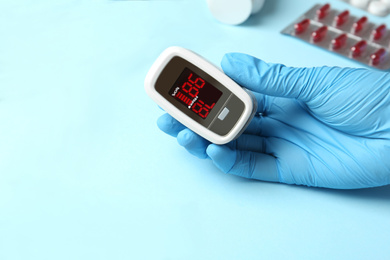 Doctor in latex gloves holding fingertip pulse oximeter near stethoscope and pills on light blue background, closeup