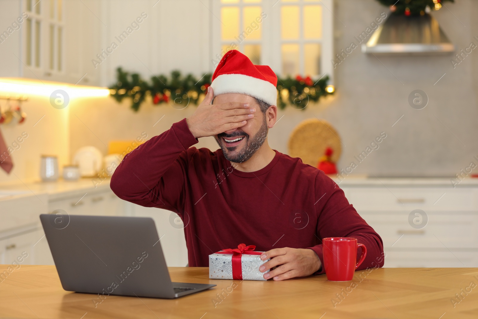 Photo of Celebrating Christmas online with exchanged by mail presents. Man in Santa hat covering eyes before opening gift box during video call on laptop at home