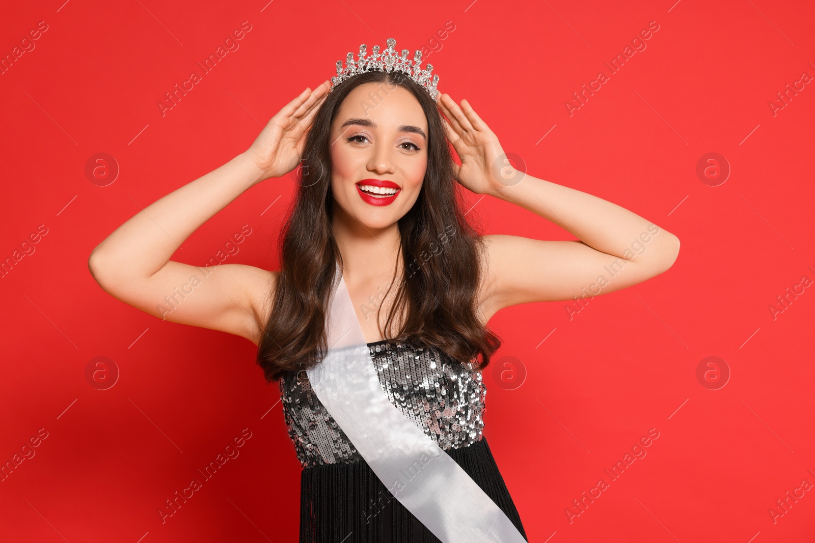 Photo of Beautiful young woman with tiara and ribbon in dress on red background. Beauty contest