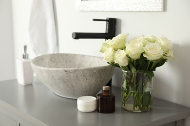 Vase with beautiful white roses and toiletries near sink in bathroom