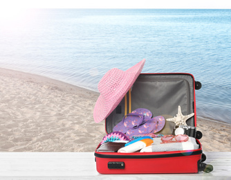 Image of Suitcase with different beach objects on wooden table near sea. Space for text