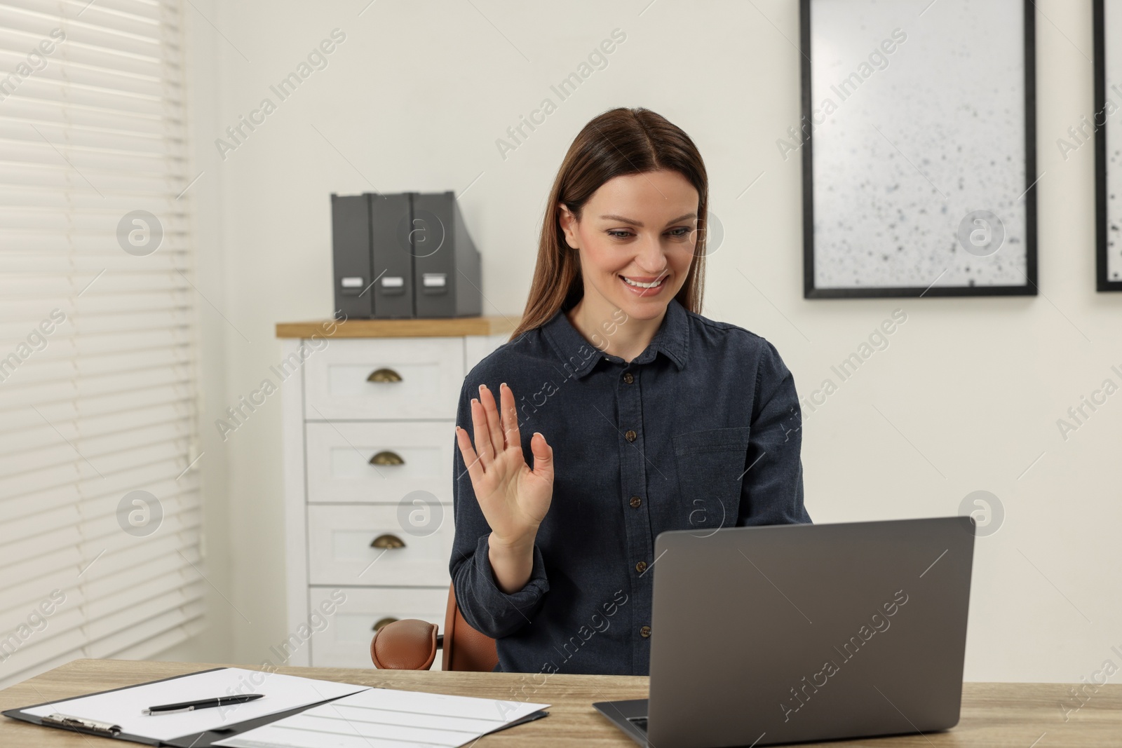 Photo of Woman waving hello during video chat via laptop at wooden table in office