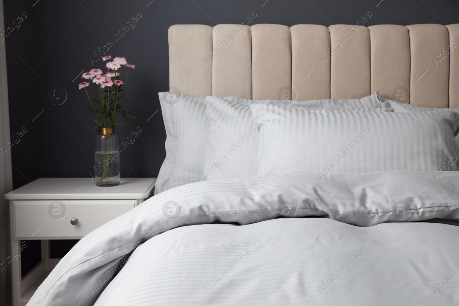 Photo of Nightstand and flowers near comfortable bed with soft pillows indoors