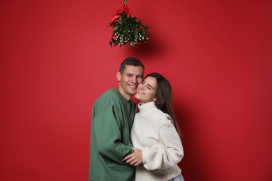 Photo of Happy couple standing under mistletoe bunch on red background